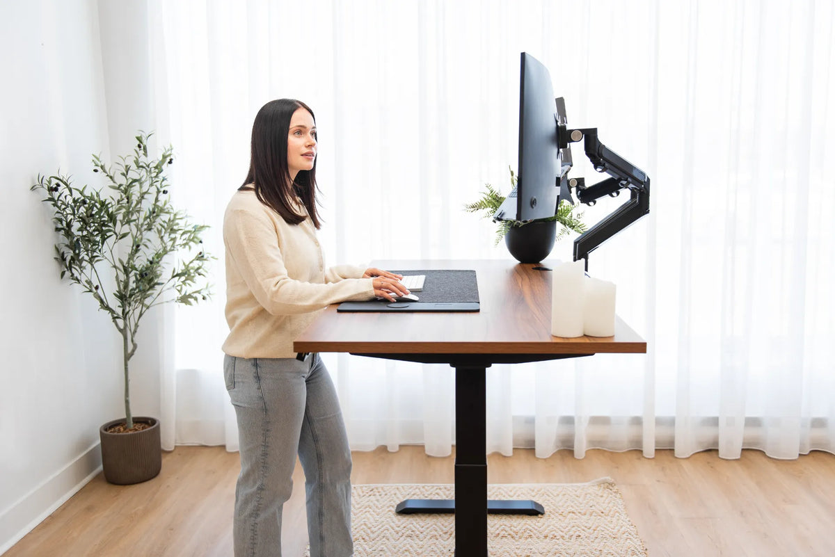 Person standing at a wood standing desk with two monitors on black adjustable arms in a bright office