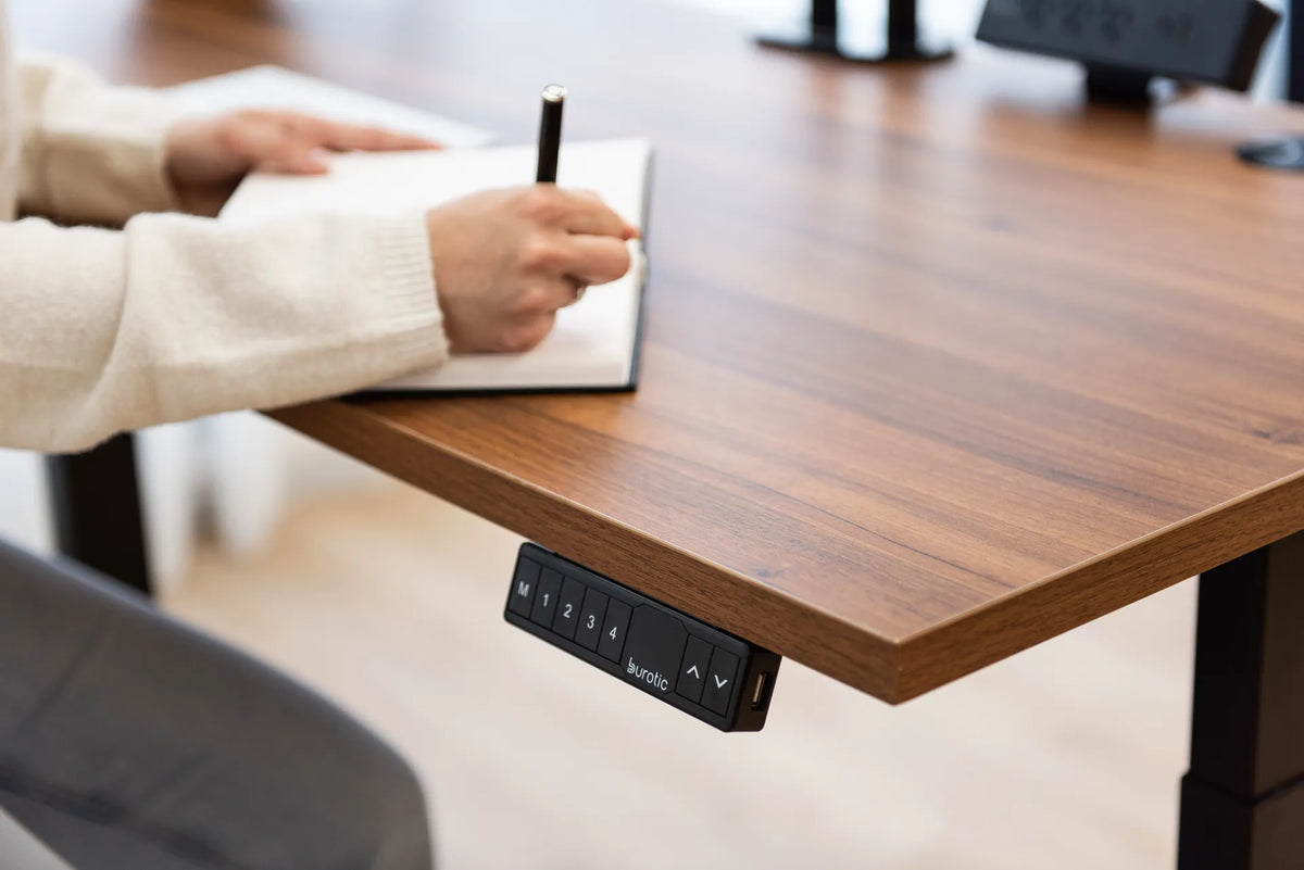 Person writing in an open notebook on a wood standing desk with a black memory controller below the desktop