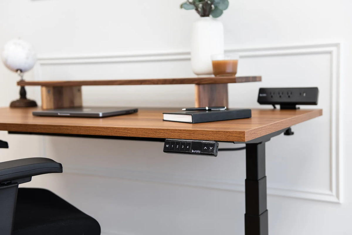 Brown wood standing desk top with a black memory controller, monitor shelf, notebook, closed laptop and desktop power bar