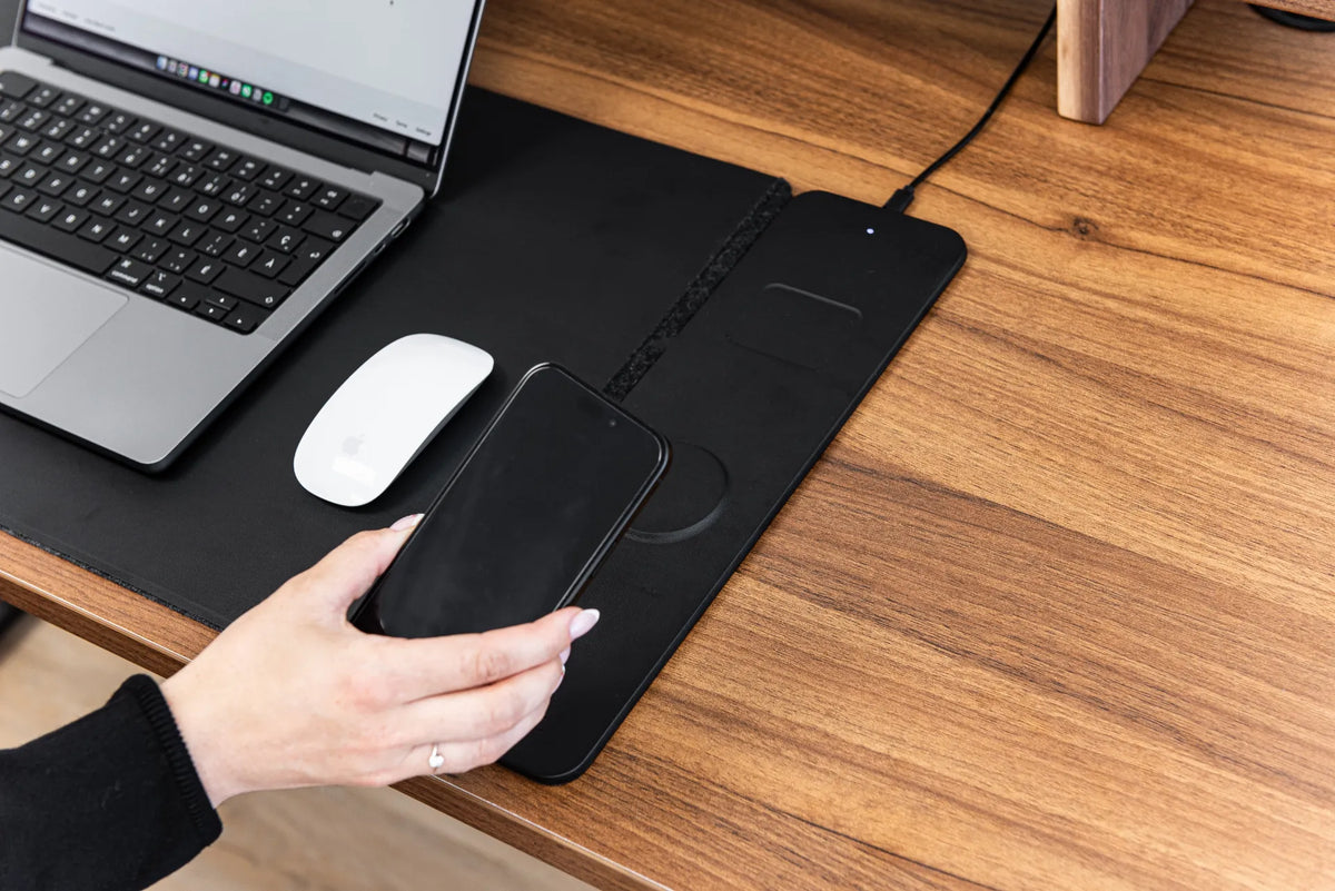 Hand positioning a black smartphone over the charging area of a black desk pad beside a laptop and white mouse on a wood desk