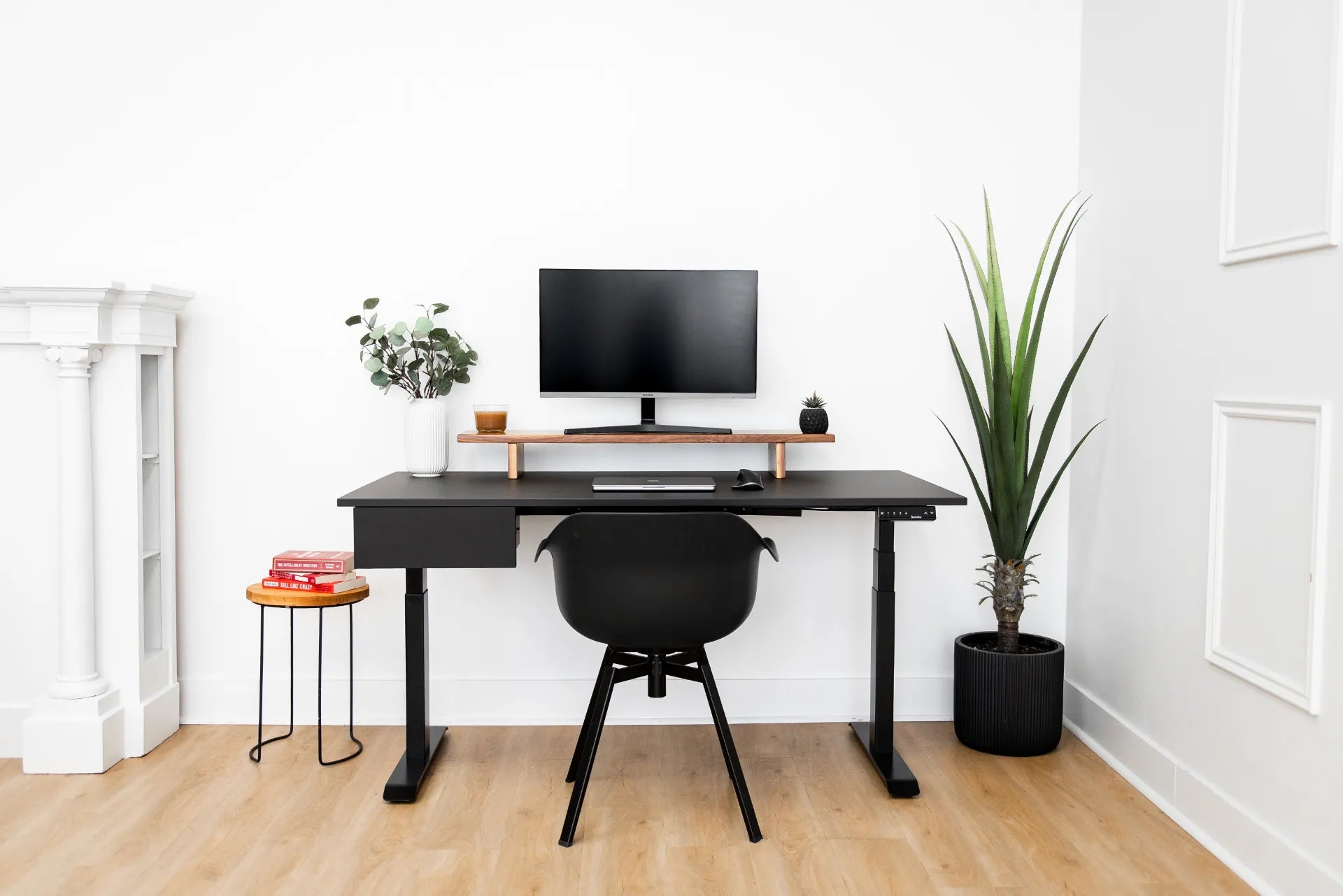 Full black adjustable desk frame and desktop in a bright white office with a monitor, shelf, chair and plants