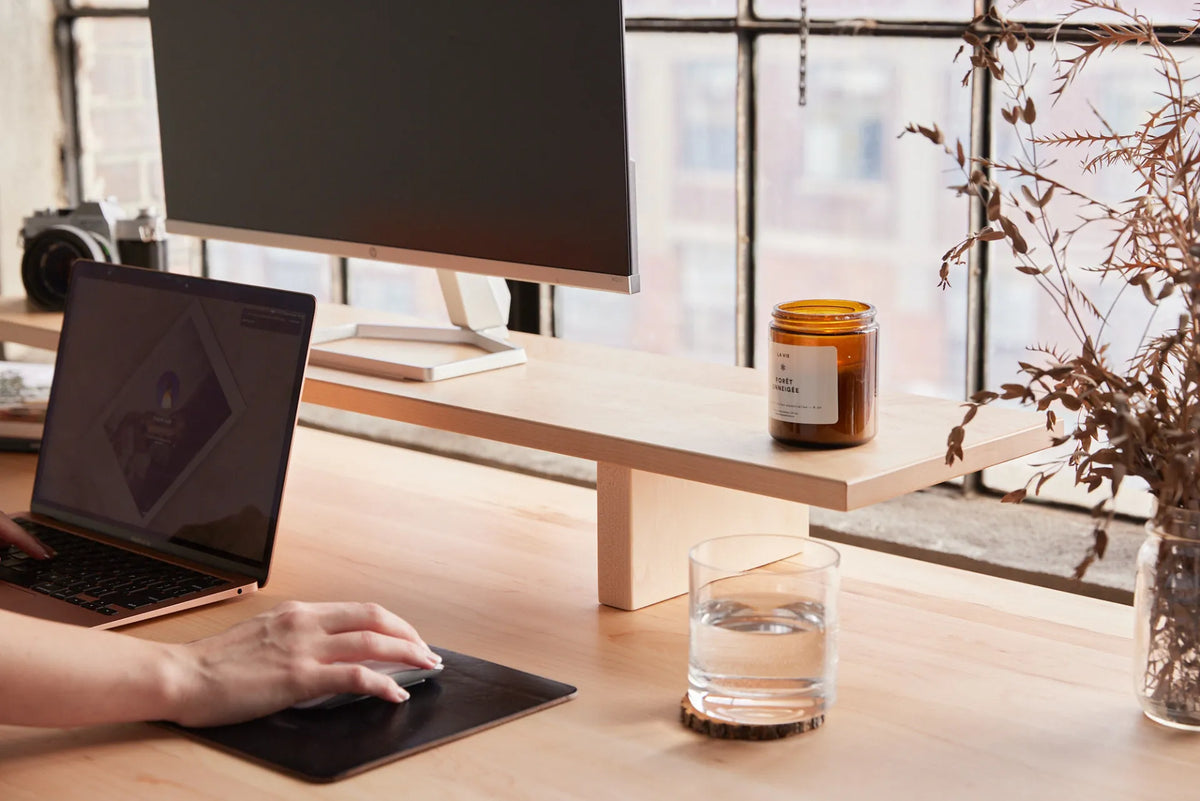 Large monitor on a light wood riser above a laptop workstation beside industrial windows