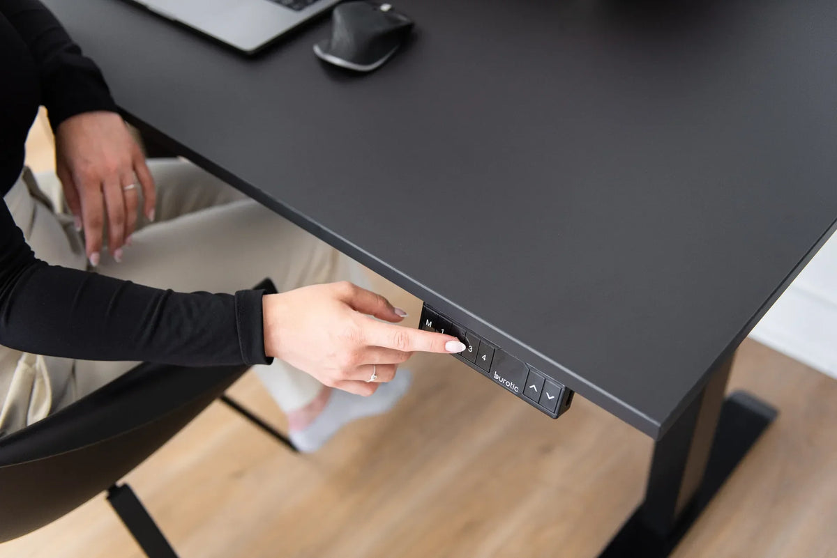 Hand pressing a memory preset on a black Burotic standing desk controller beside a laptop and mouse