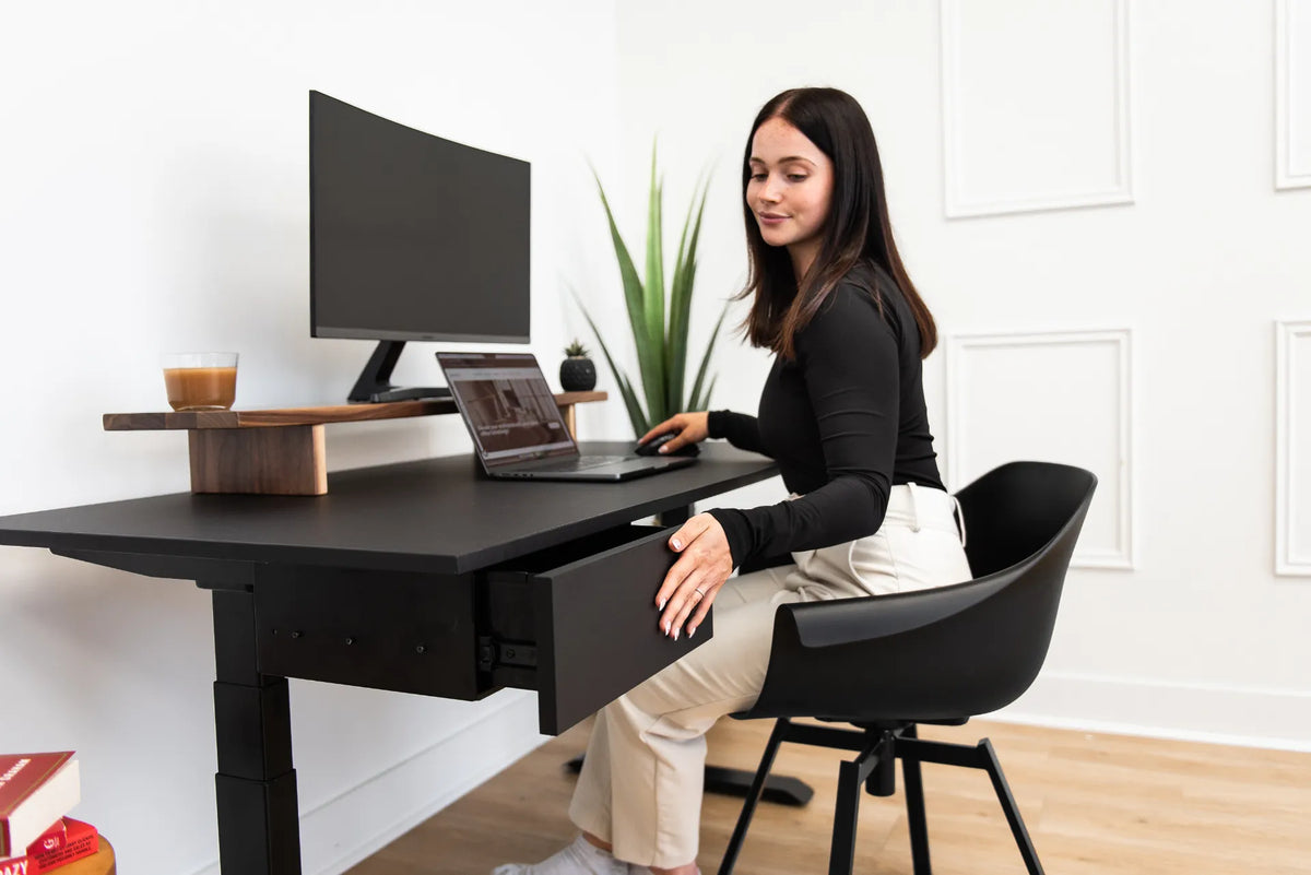 Woman seated in a black chair at a black desk while opening an under-desk drawer in a bright office