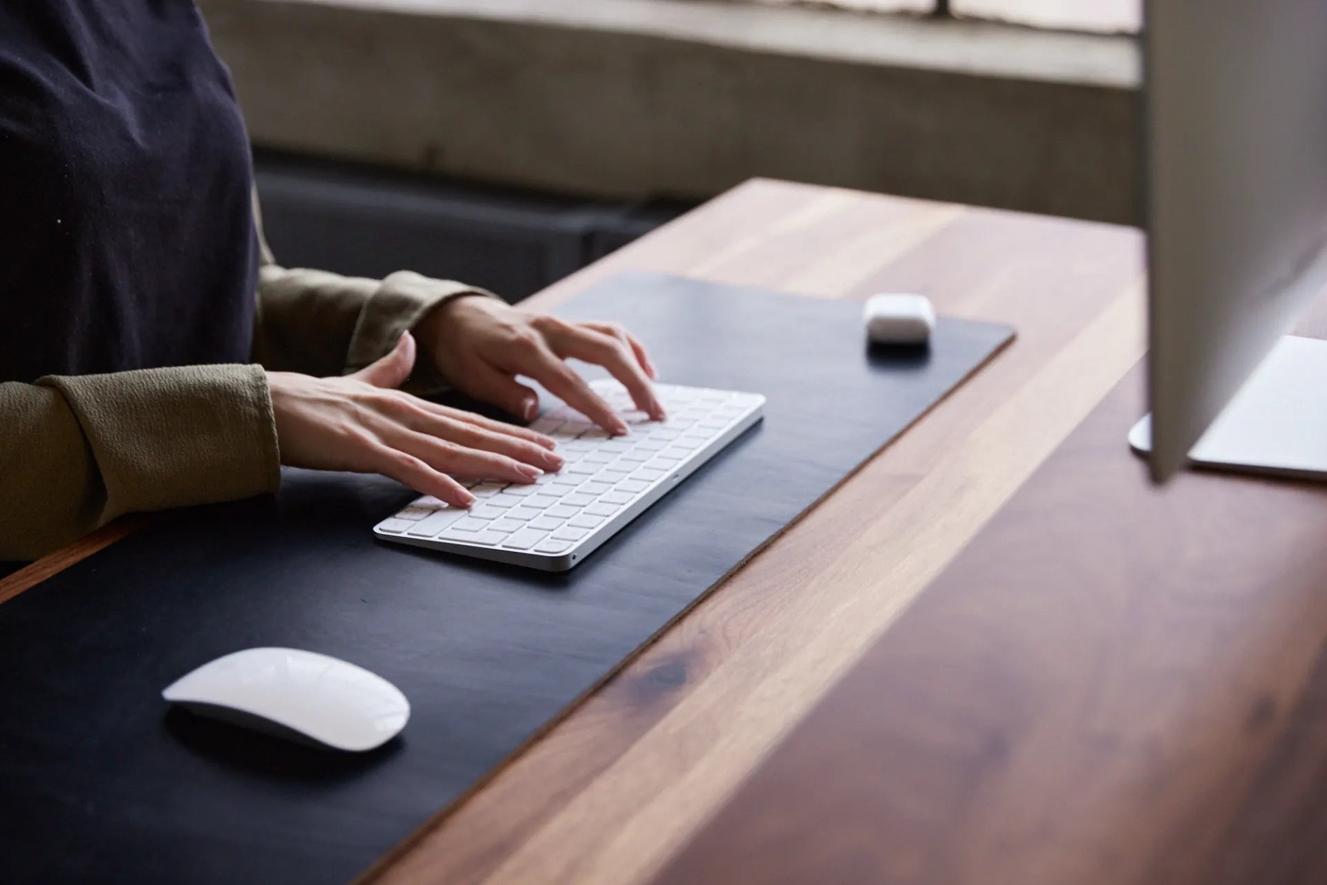 Hands typing on a compact white keyboard beside a mouse on a black desk mat and wood desktop