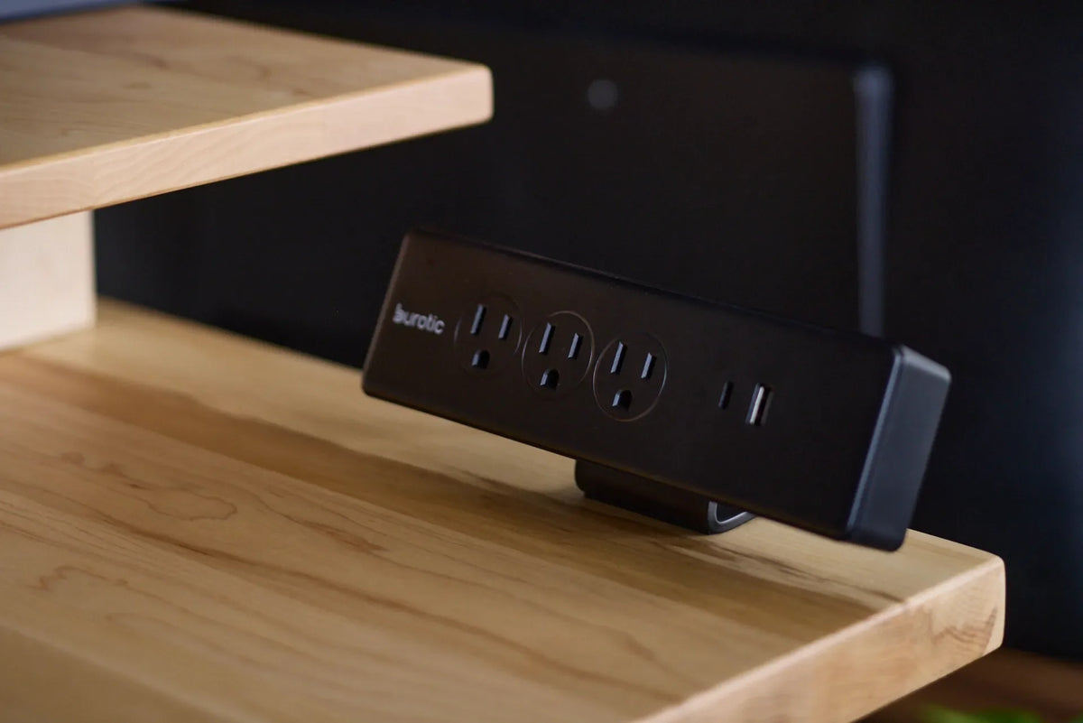 Black Burotic desktop power bar with three outlets, USB-C and USB-A ports on a light wood monitor stand