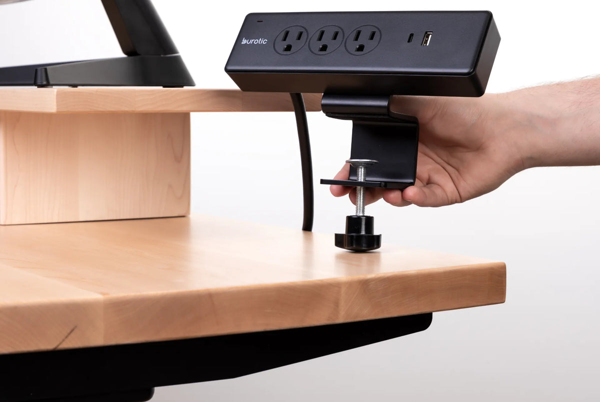 Hand tightening a black clamp-on Burotic power bar to the edge of a light wood desk
