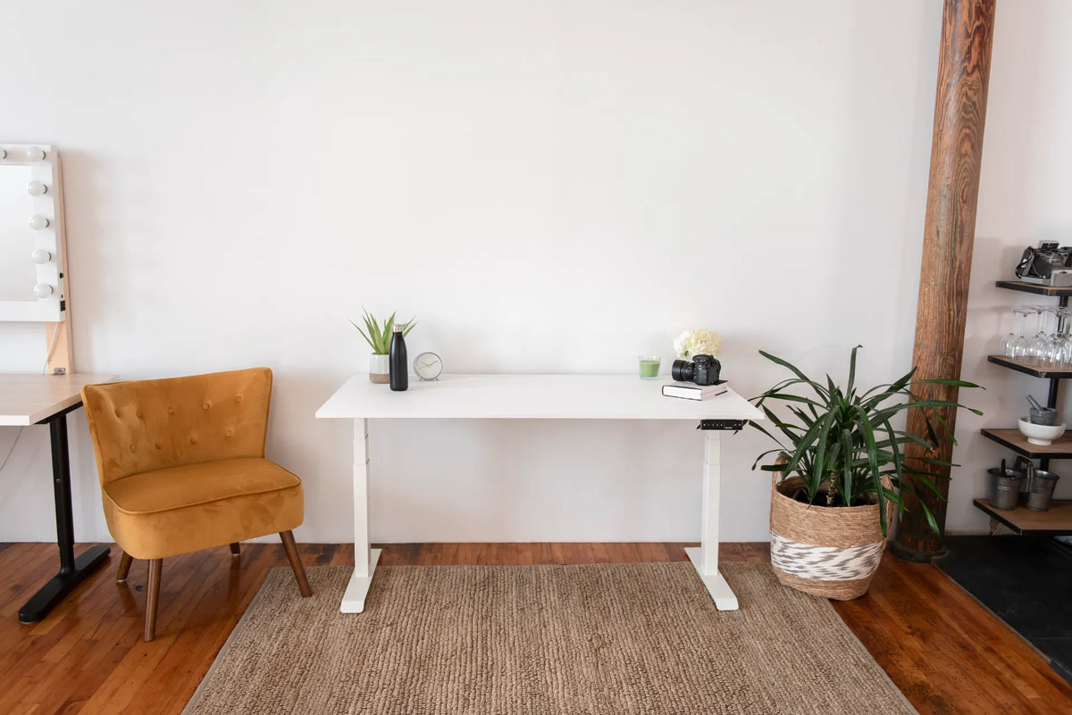 Compact white standing desk with a monitor, small plant and lamp in a bright room with a lounge chair