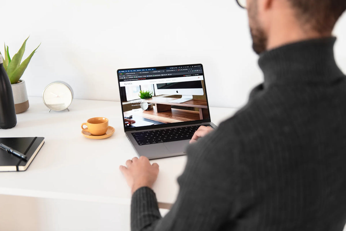 Person browsing the Burotic website on a laptop at a white desk with an orange cup, clock and plants