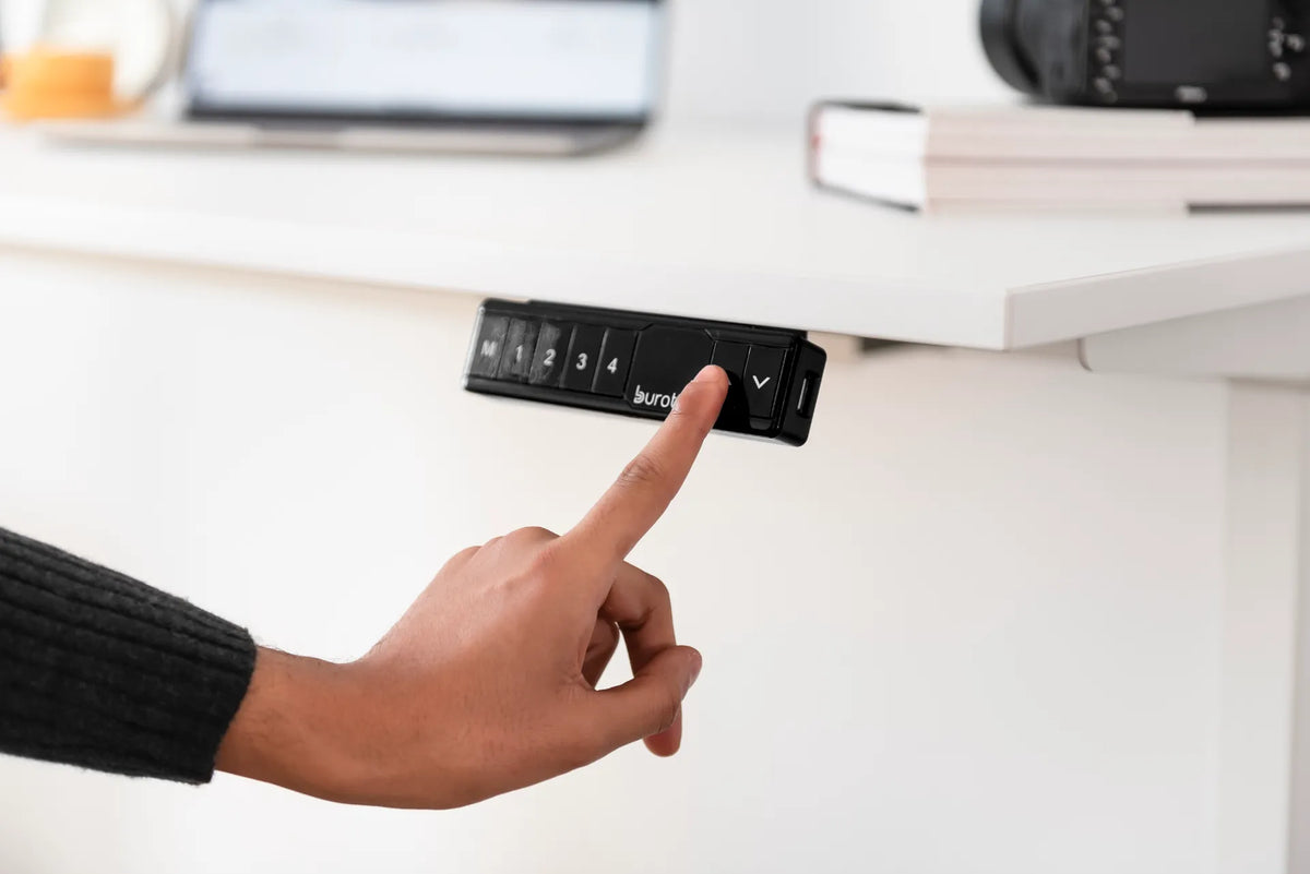 Hand pressing the up arrow on a black Burotic memory controller mounted beneath a white standing desk
