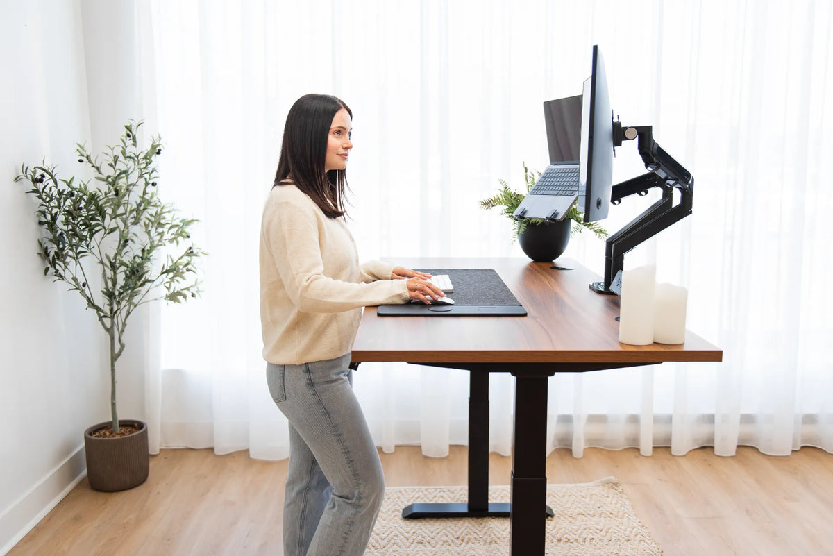 Woman standing at a walnut-tone adjustable desk with a curved monitor and laptop mounted on separate black arms