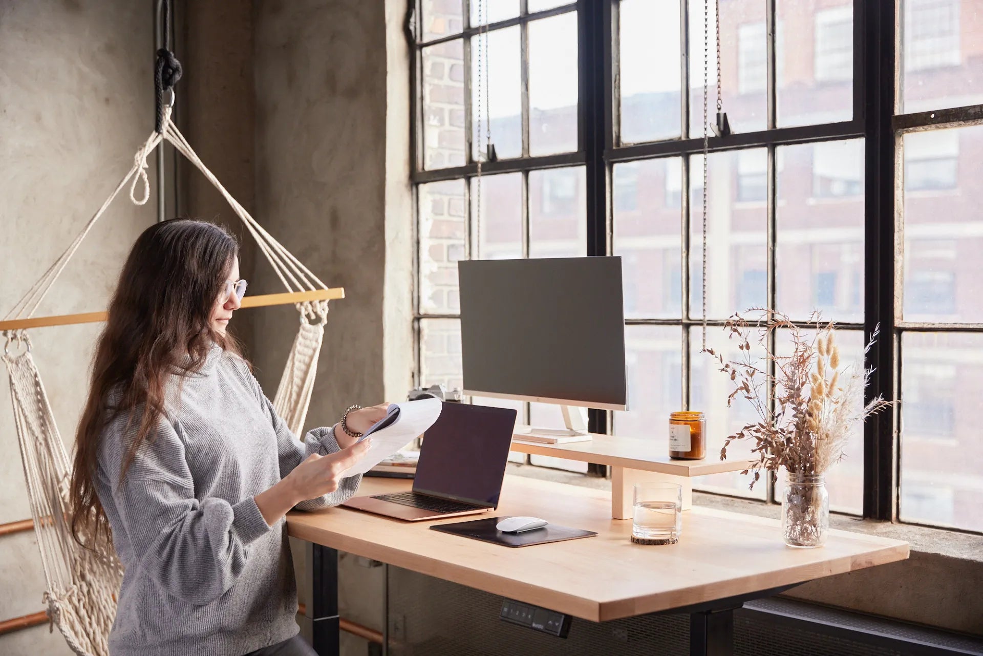 Person standing at a light wood desk with a laptop, monitor and large industrial windows