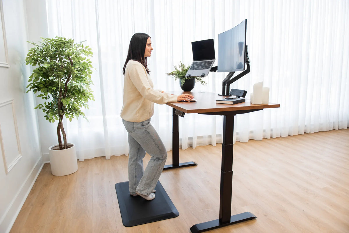 Person standing at a wood adjustable desk with a laptop to the left of a curved external monitor on black arms