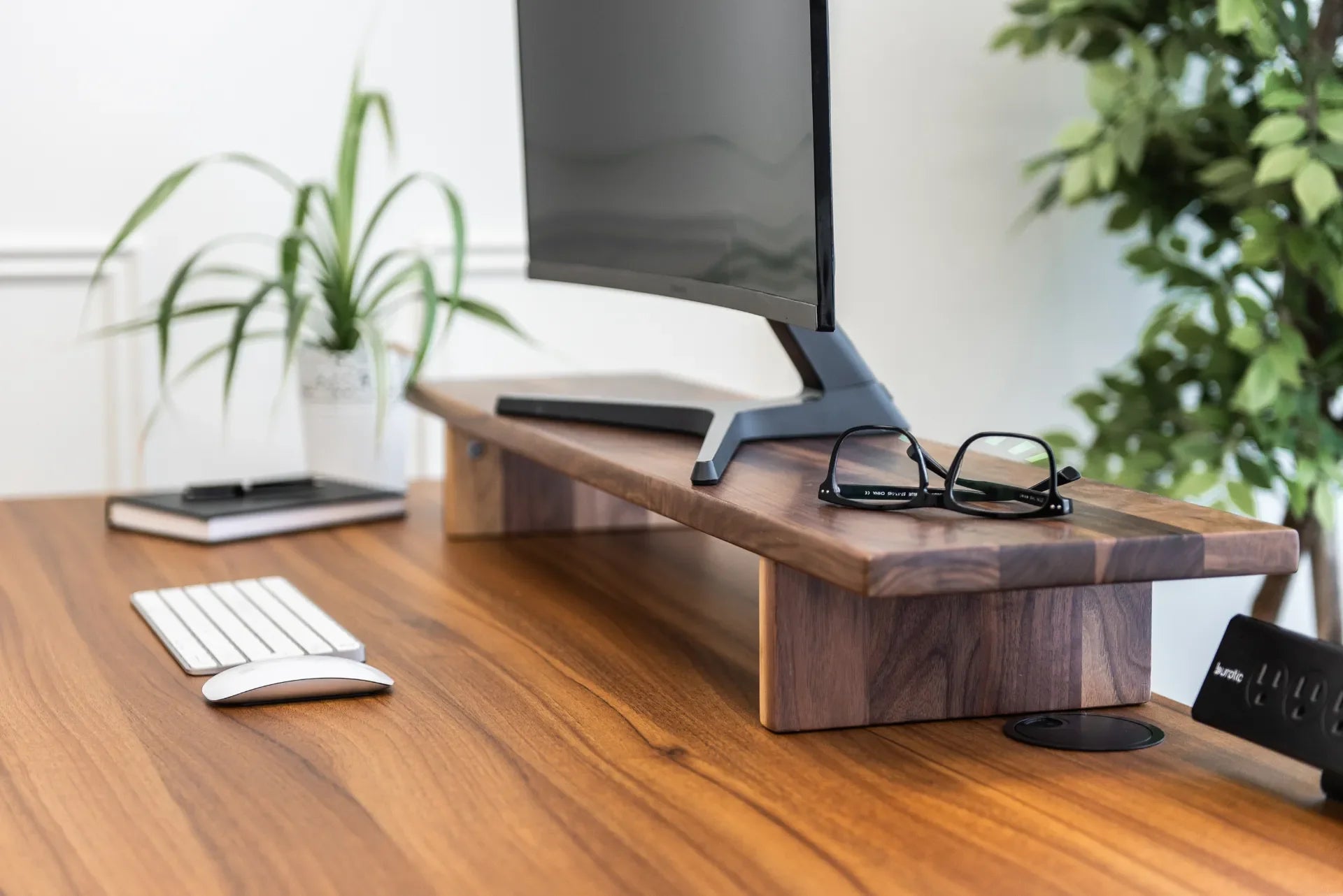 Close view of a monitor on a walnut Burotic stand with a keyboard, mouse, glasses and plants