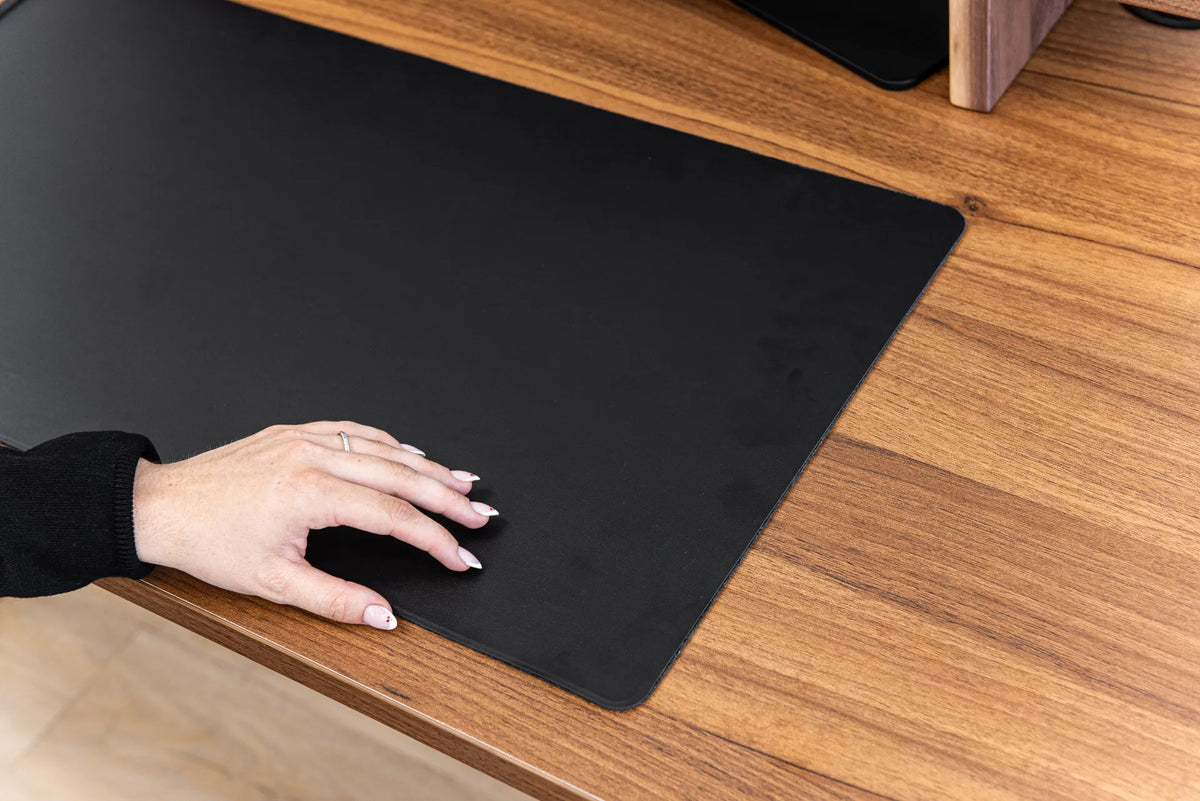Black desk pad with a keyboard and mouse on a warm wood desktop, shown in close-up