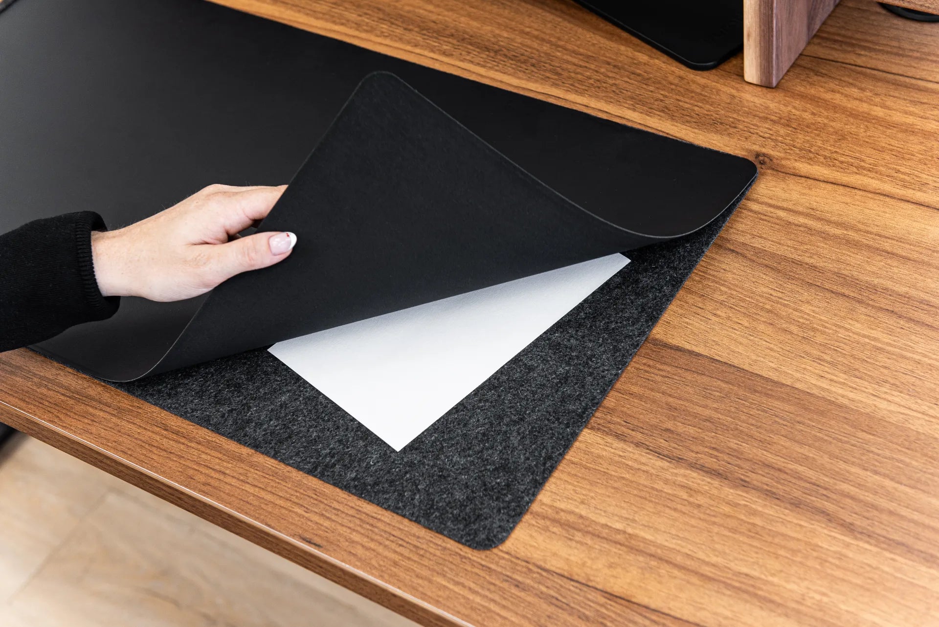 Hand lifting the black top layer of a desk pad to reveal felt backing and stored papers on a walnut desktop