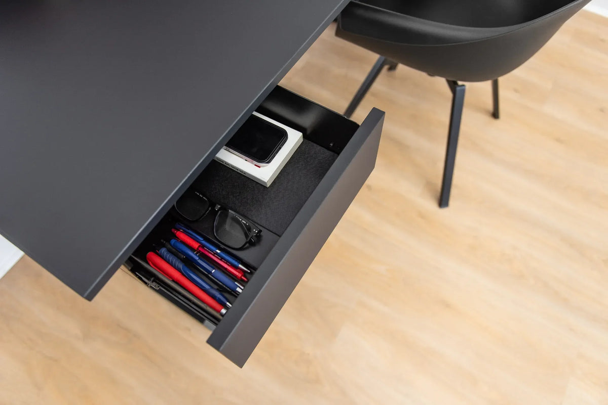 Open black under-desk drawer holding pens, glasses and a phone beneath a black desktop