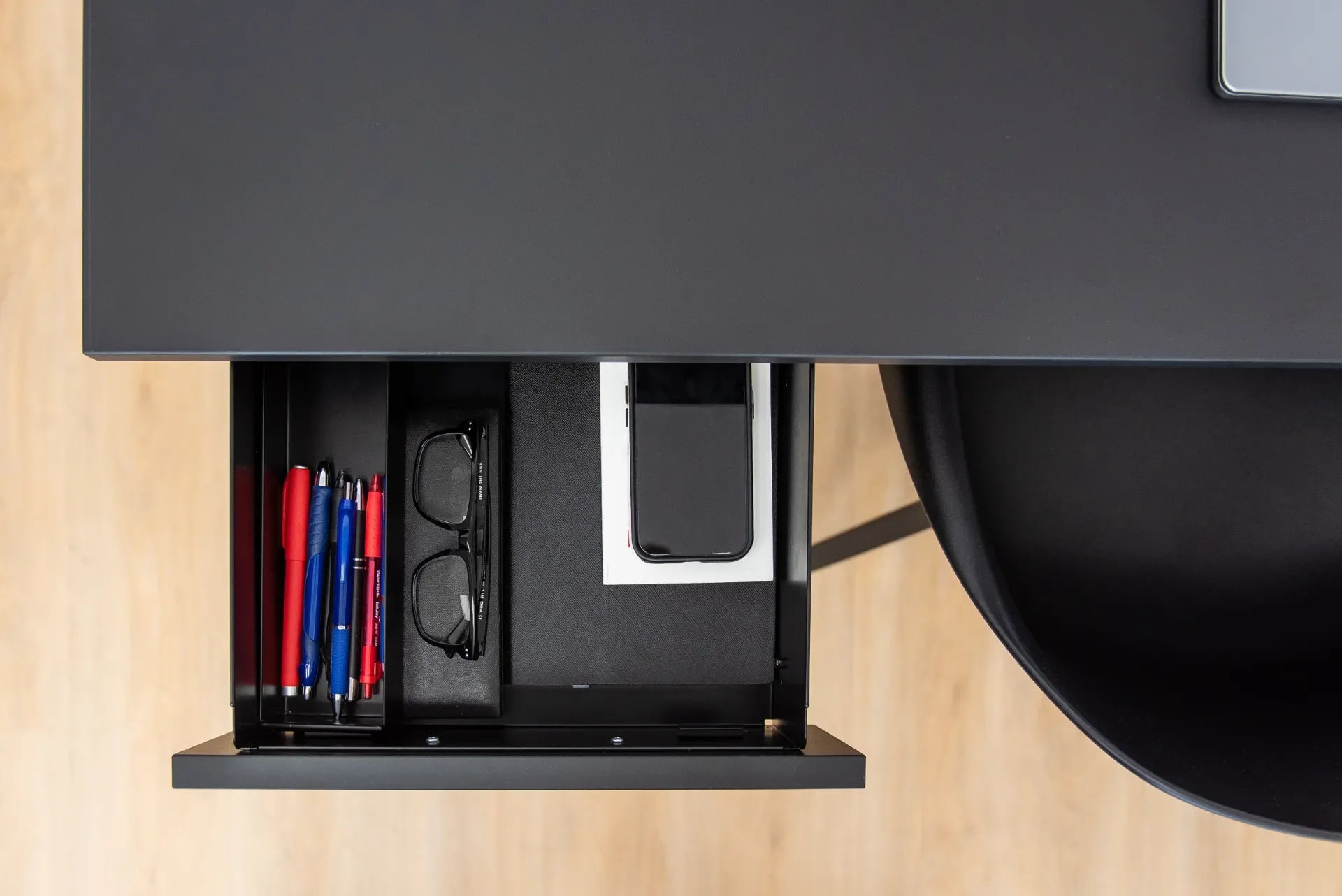 Open black desk drawer organized with pens, glasses, a notebook and a smartphone