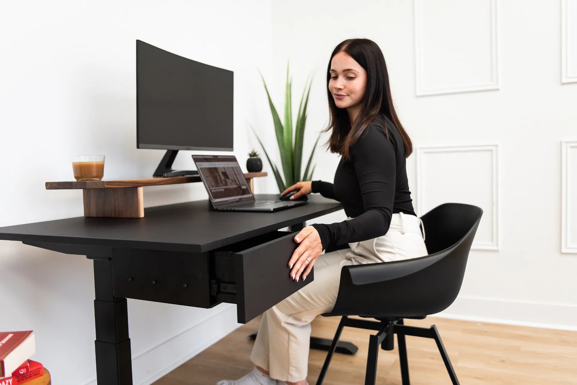 A person seated at a black desk with a drawer, monitor stand and organized work surface