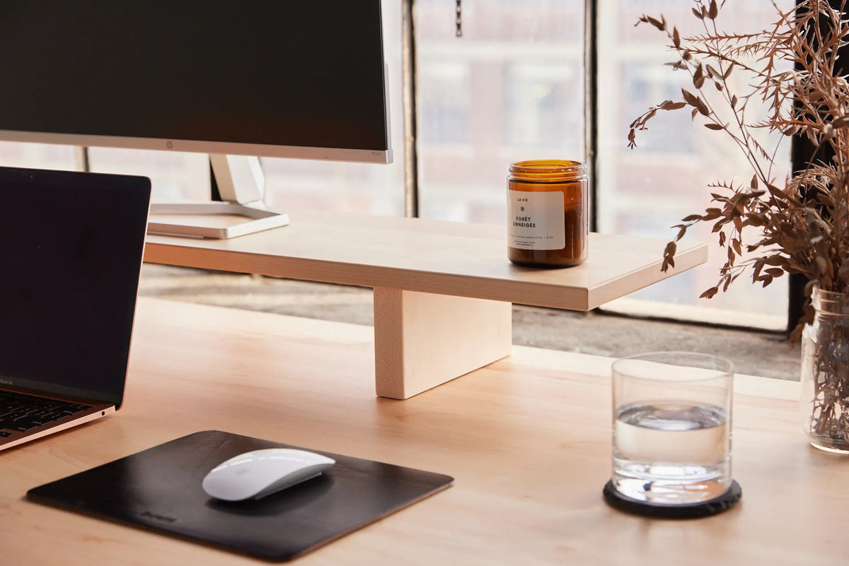 Glass of water on a coaster beside a white mouse, black mouse pad, laptop and monitor stand on a light wood desk
