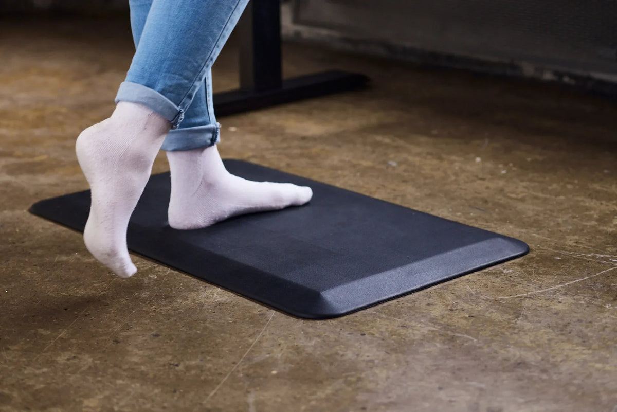 Close-up of socked feet shifting position on a black anti-fatigue mat beneath a standing desk on a concrete floor