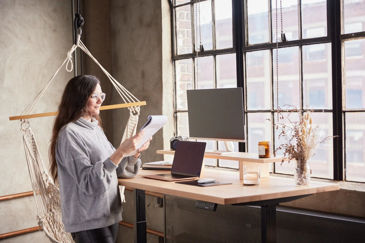 Woman standing at a light wood adjustable desk and reviewing paper documents beside a laptop, monitor and monitor stand