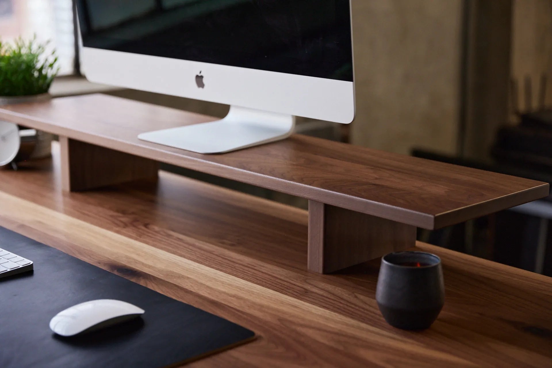 Dark wood monitor stand supporting a desktop display on a wood desk beside a mug and mouse