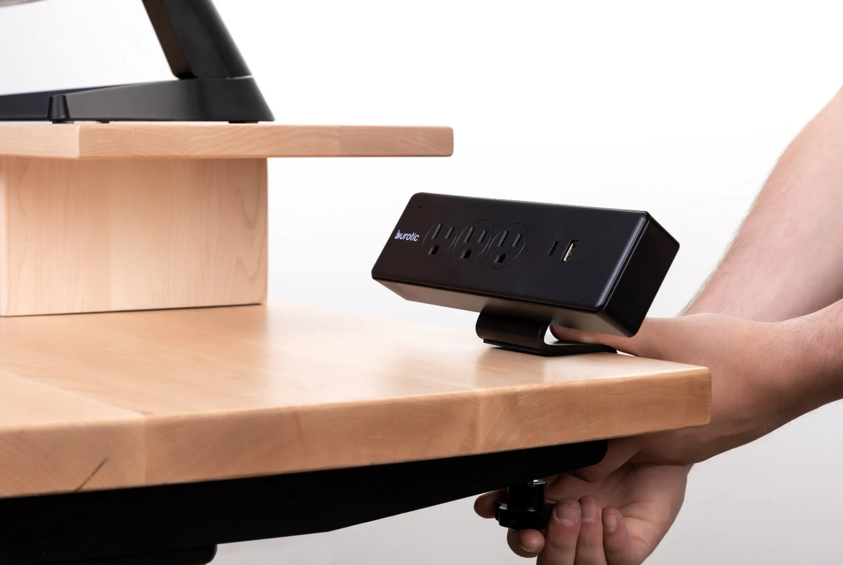 Hands tightening the clamp of a black Burotic power bar to the edge of a light wood desktop