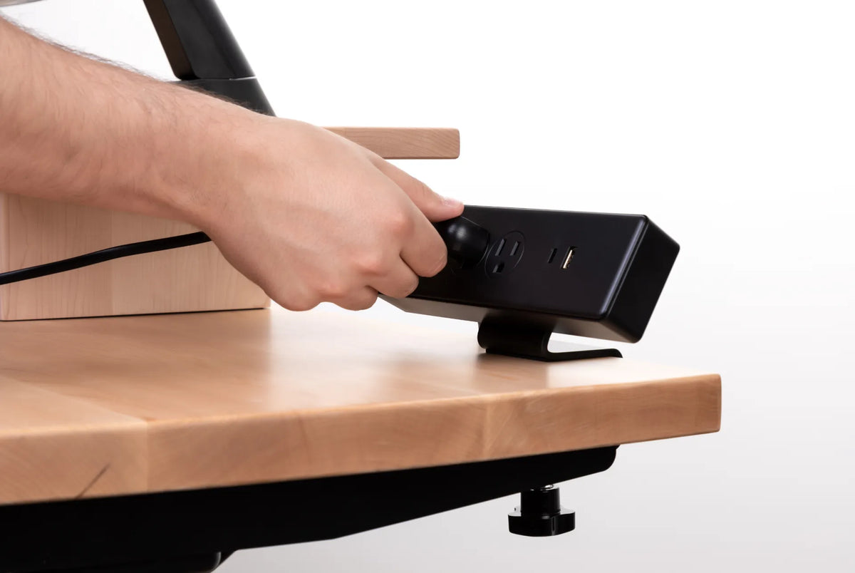 Hands connecting a black power bar on a light wood desk shelf with a clamp mount visible below