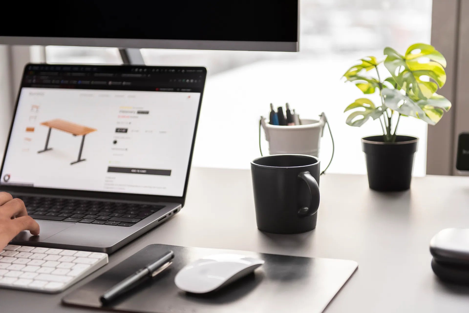 Minimalist black desk with laptop, monitor, white keyboard, mouse, notebook, mug and plant