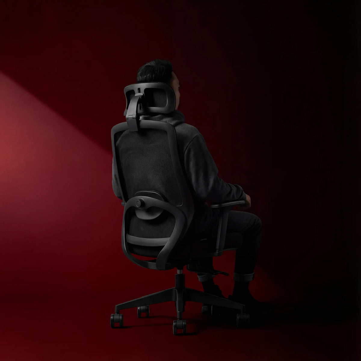 Person seated in a black mesh ergonomic chair with headrest against a dark red studio background