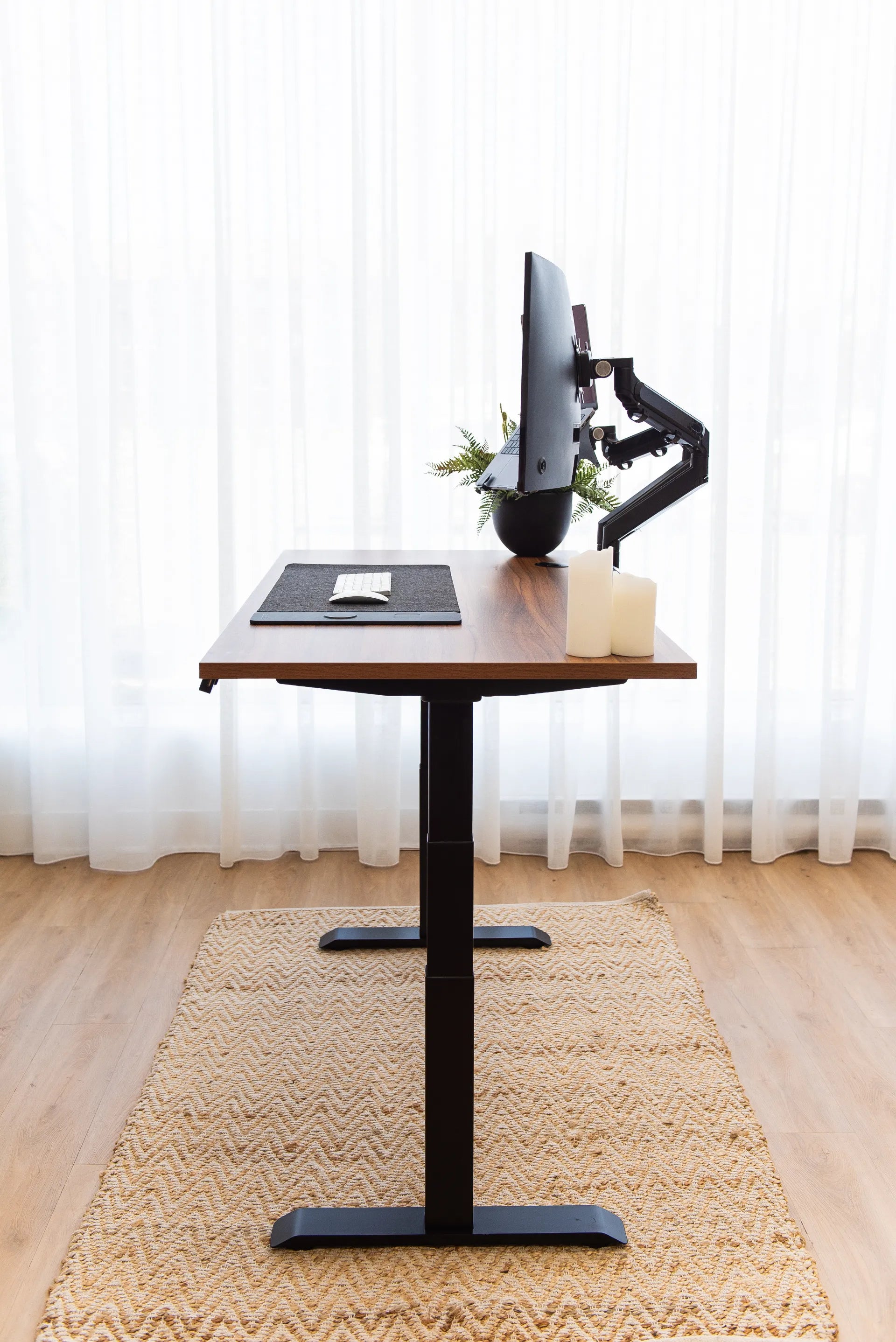 Side view of a wood standing desk with black frame, monitor arms, keyboard and floor rug