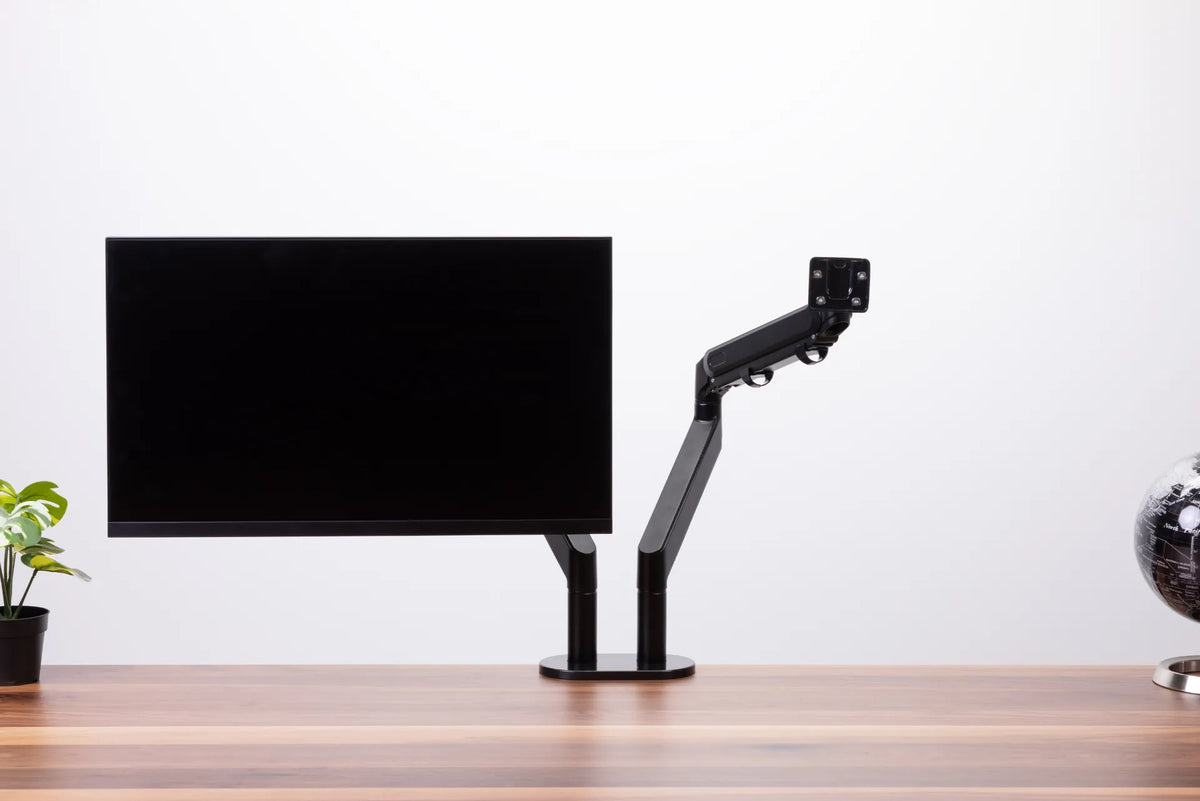 Black monitor arm demonstration with one screen attached and a second mounting arm visible above a wood desk