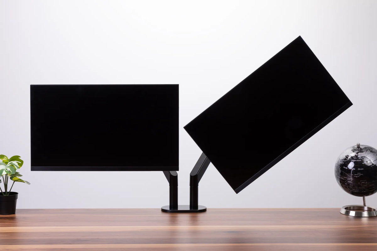 Two black monitors of similar size on a black dual monitor arm above a wood desk, with one screen angled inward