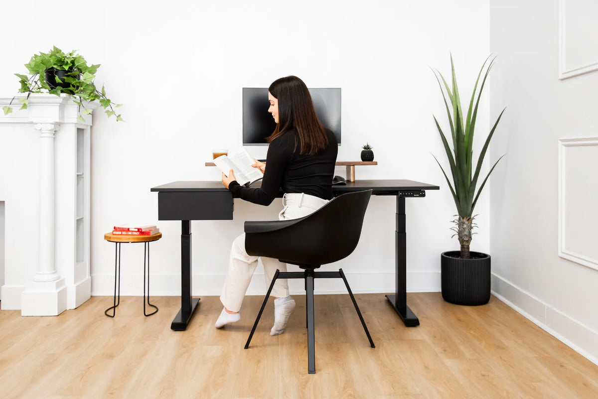 Woman reading at a black adjustable desk with black top, monitor, under-desk drawer and plants in a bright room