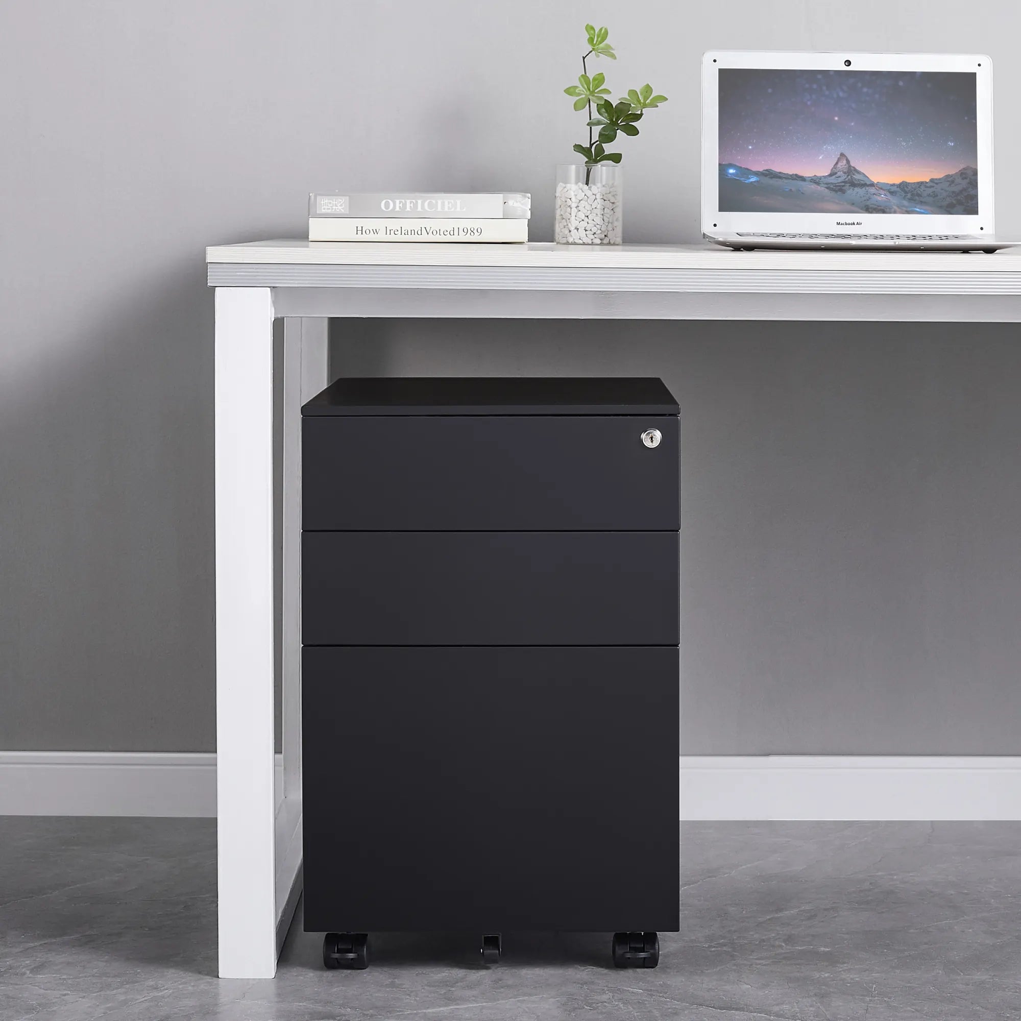 Black three-drawer filing cabinet next to a white desk with a laptop and books on a gray wall background.